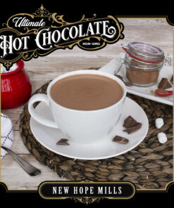 An image showing prepared Ultimate Hot Chocolate in a mug, with bits of chocolate, marshmallows, and kitchenware around the mug. Image contains text 
