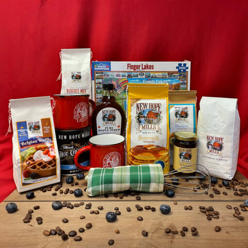 Product display of 'Breakfast with the Crew' collection, featuring a 2lb bag of Buttermilk pancake mix, a 1.5lb bag of biscuit mix, a 1lb bag of coffee, a 1lb canister of Original Ultimate Hot Chocolate mix, a 1.5lb bag of Belgian Waffle mix, an 8oz bag of Blueberry pancake mix, two ceramic mugs, a wire whisk, a tea towel, and a 1,000-piece jigsaw puzzle, all arranged on a wooden surface, with coffee beans and blueberries strewn on the wooden surface.