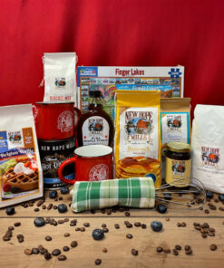 Product display of 'Breakfast with the Crew' collection, featuring a 2lb bag of Buttermilk pancake mix, a 1.5lb bag of biscuit mix, a 1lb bag of coffee, a 1lb canister of Original Ultimate Hot Chocolate mix, a 1.5lb bag of Belgian Waffle mix, an 8oz bag of Blueberry pancake mix, two ceramic mugs, a wire whisk, a tea towel, and a 1,000-piece jigsaw puzzle, all arranged on a wooden surface, with coffee beans and blueberries strewn on the wooden surface.