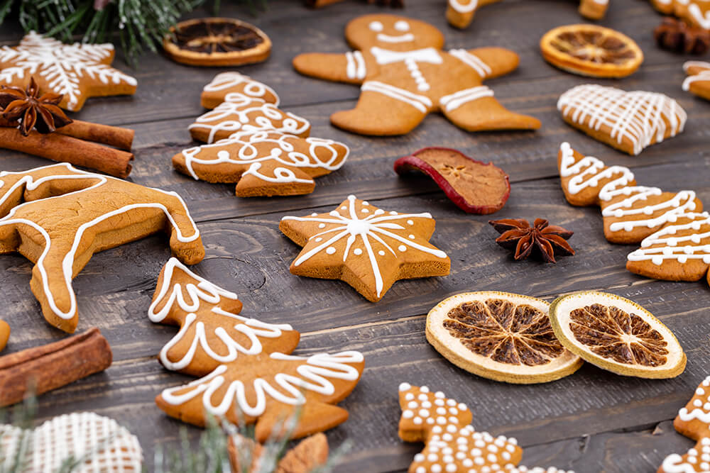 An array of gingerbread cookies with white icing decoration, in various Christmas shapes like trees, stars, and gingerbread men, arranged on a dark wooden surface. Accompanied by dried orange slices, cinnamon sticks, and star anise, evoking a festive and aromatic atmosphere.