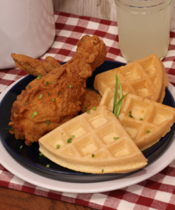 image of chicken and waffles on a plate dress with some green chives. on a table with a glass of lemonade and a gingham checkered table cover