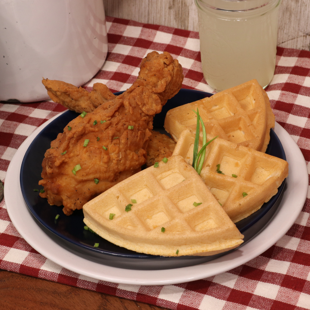 image of chicken and waffles on a plate dress with some green chives. on a table with a glass of lemonade and a gingham checkered table cover