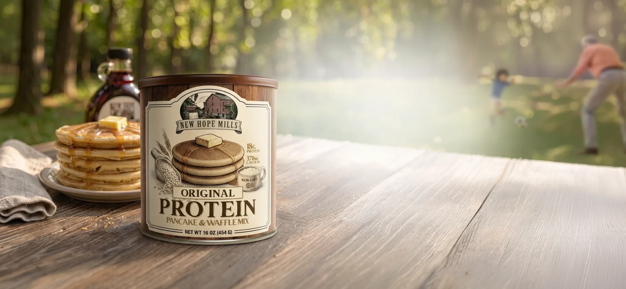 Scene of a table surface, with Protein Pancake Mix canister and plated pancakes and bottle of syrup on the table behind the canister. The background is a blurred out yard with a grandson and grandfather playing soccer together.
